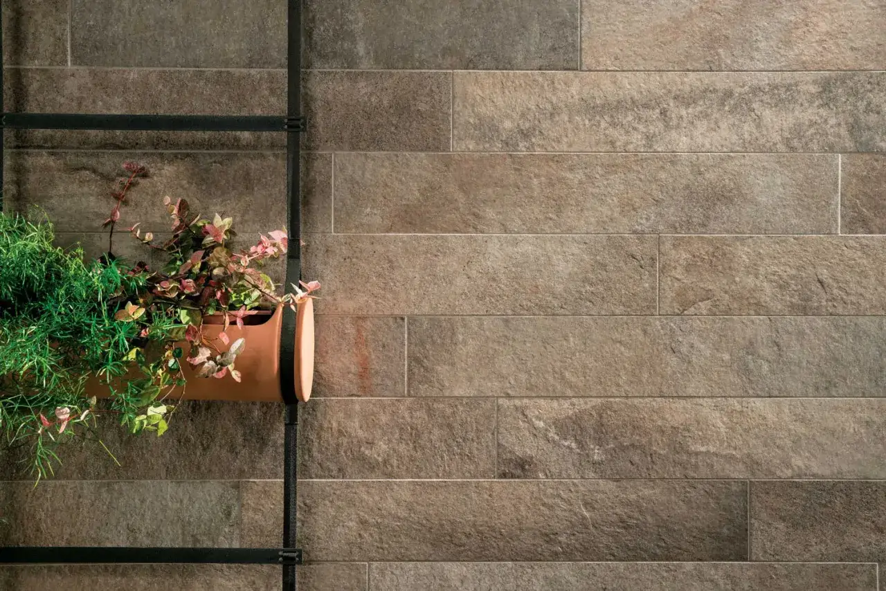 Un vaso di terracotta con piante verdi e rosa è appeso a una struttura metallica contro un pavimento non rettificato effetto pietra.