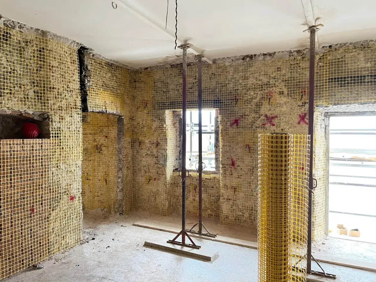 Interno di un edificio in ristrutturazione con pareti ricoperte da rete gialla per intonaco armato. Puntelli metallici sostengono il soffitto.