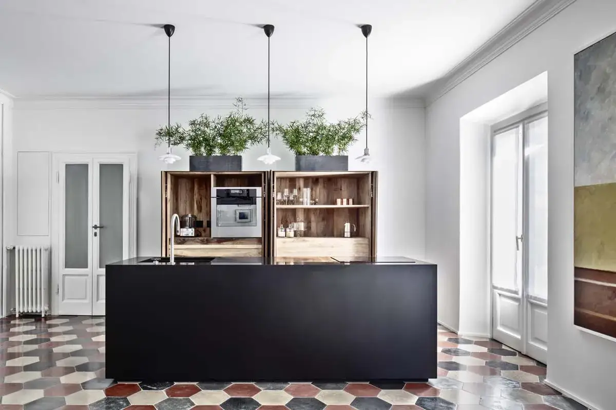 Moderna cucina con isola nera, pensili in legno con piante e forno a vista, su un pavimento con colorate cementine esagonali.