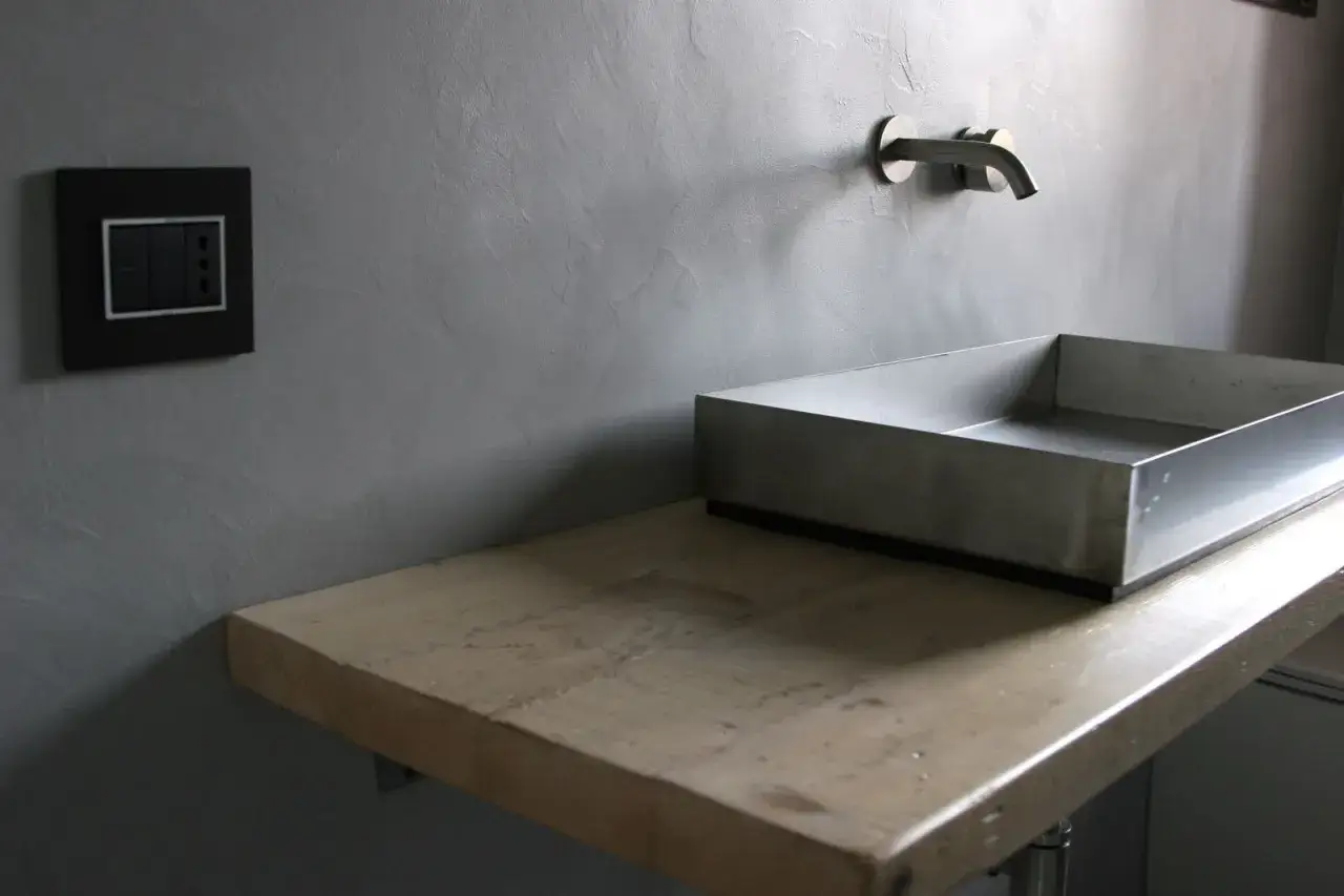 Lavandino in metallo su mensola di legno grezzo, con rubinetto a muro. Un'idea moderna per coprire piastrelle in bagno.