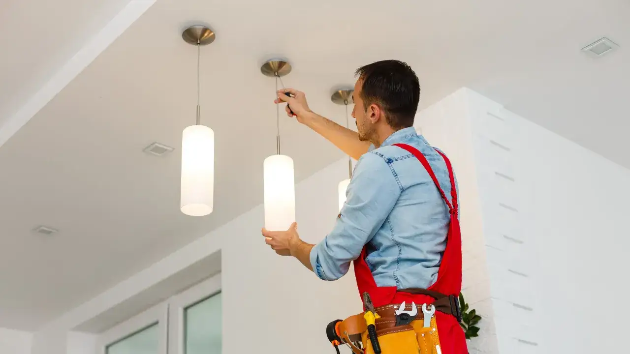 Elettricista al lavoro per il montaggio lampadario a sospensione. L'uomo con tuta rossa e cintura porta attrezzi fissa le luci al soffitto.