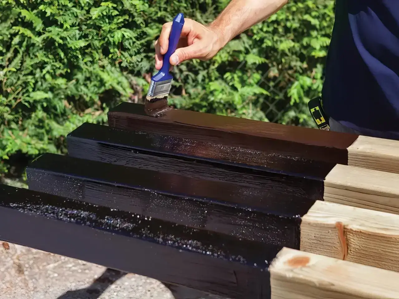 Mano che applica vernice scura su listelli di legno per impermeabilizzare il legno fai da te.
