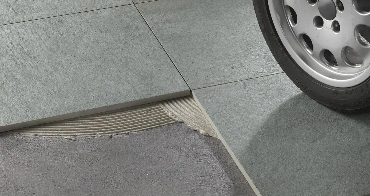 Dettaglio di piastrelle grigie con colla visibile sotto una sollevata. Lo spessore della colla piastrelle è evidente, pronto per l'installazione.