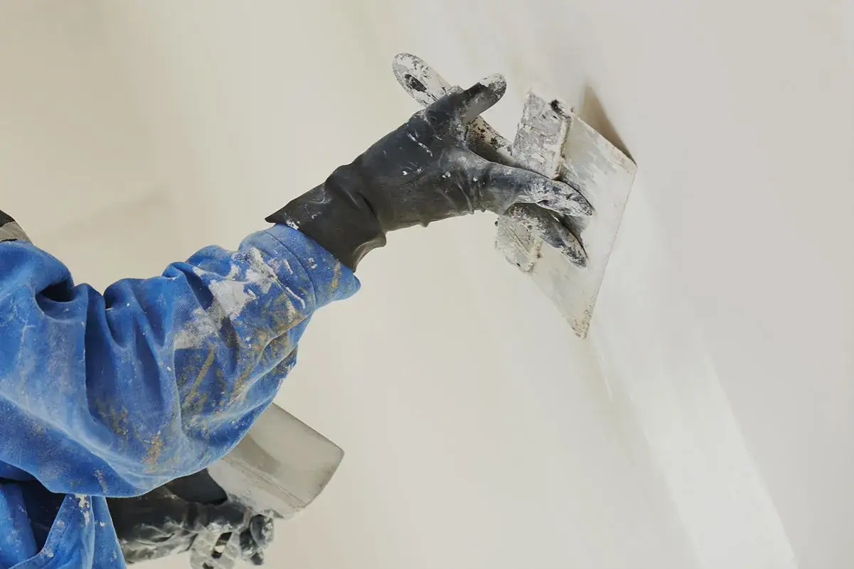 Mano guantata stende rasante fino per interni su una parete, creando una finitura liscia e uniforme.