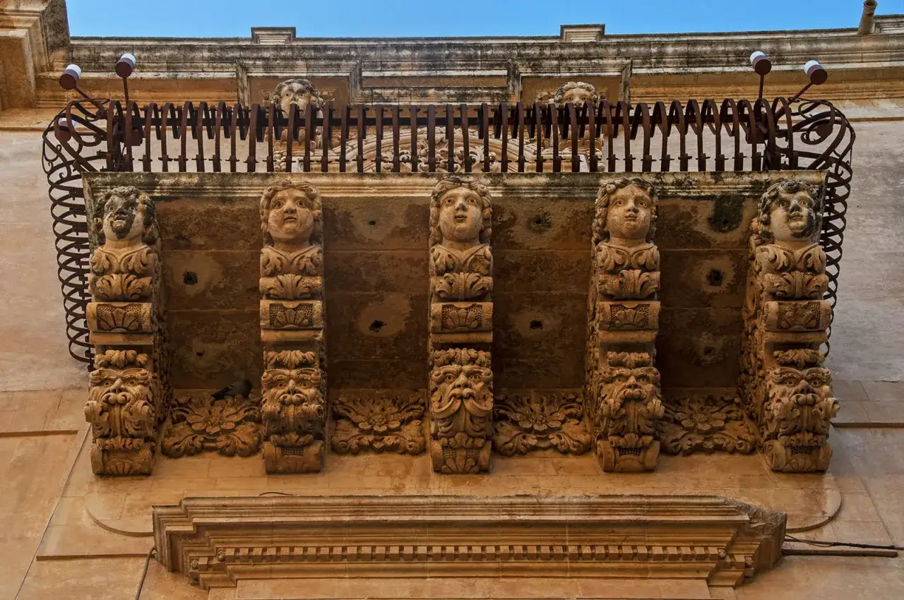 Balcone barocco con mensole scolpite a forma di volti grotteschi e putti, tipico della pietra di Noto.
