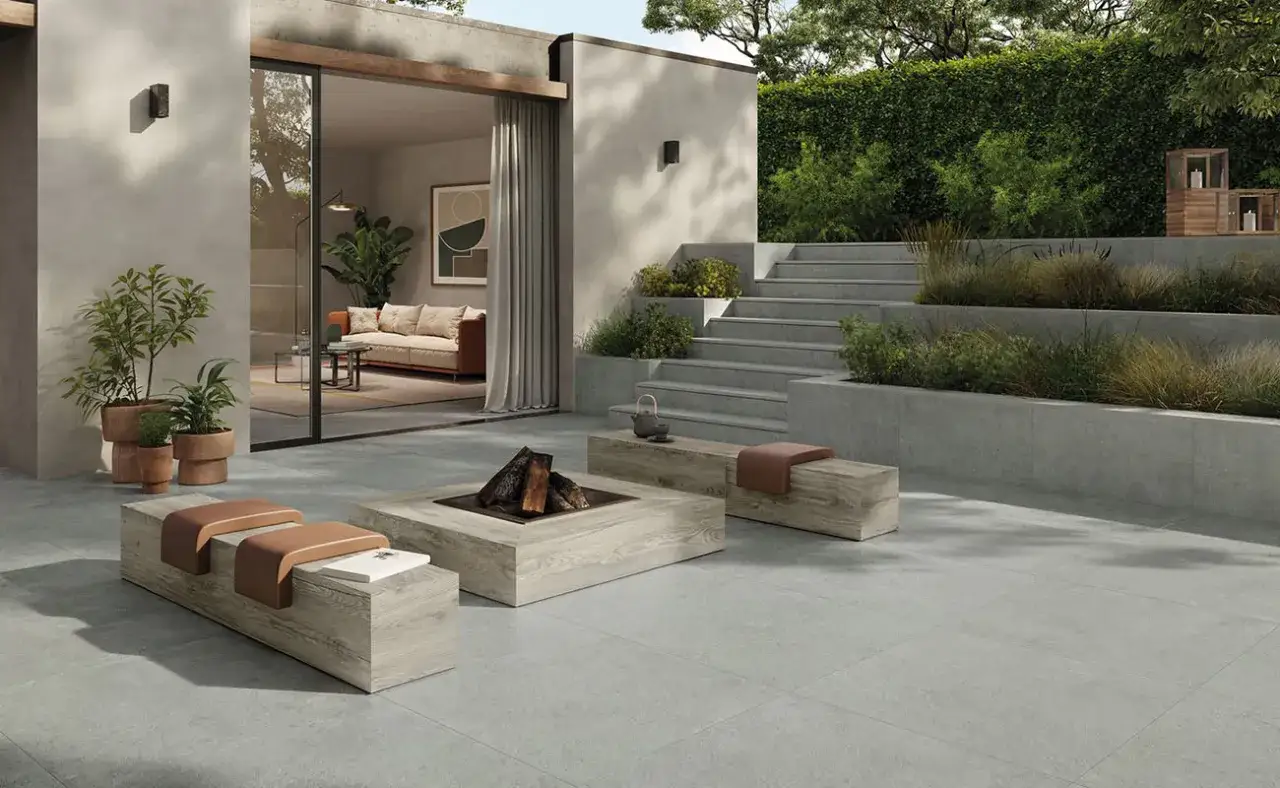 Un'elegante pavimentazione esterna moderna con un focolare centrale, panche in legno e un'area salotto interna visibile attraverso una grande vetrata.