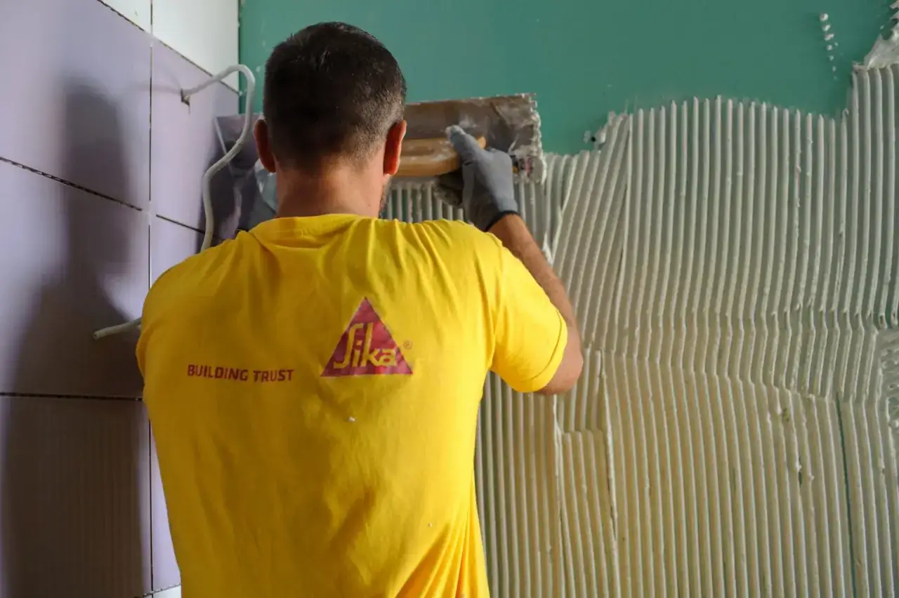 Operaio con maglia gialla Sika applica colla per la partenza posa piastrelle a muro.